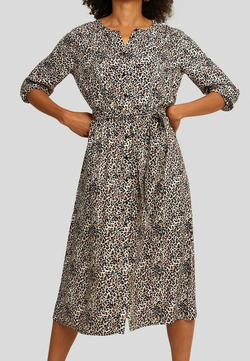 Femme portant une robe léopard mi-longue avec des boutons et des manches trois quarts, mains posées sur les hanches, sur un fond uni.