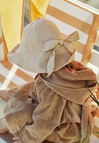 Barn iført en beige solhat med sløjfe og et brunt hættelagen, der sidder på en stribet liggestol i sollys.