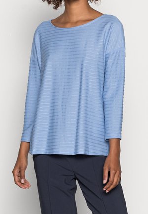 Frau trägt ein hellblaues langärmliges Oberteil mit horizontalen, strukturierten Streifen und dunkle blaue Hosen, dargestellt vom Hals bis zur Hüfte.