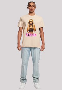 Béžové bavlněné tričko s potiskem ženy s dlouhými vlasy, rámovaným textem "britney" v růžových a bílých blokových písmenech. Džíny a bílé tenisky dokončují vzhled.