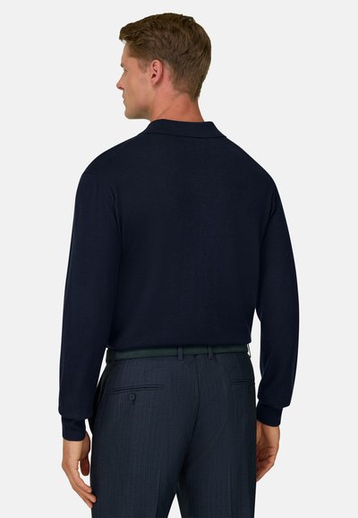 Homme de dos portant une chemise à manches longues bleu marine foncé rentrée dans un pantalon à fines rayures foncé avec une ceinture noire.
