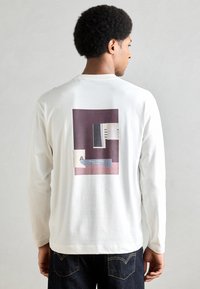 Balts krekls ar garām piedurknēm, uz muguras ir grafisks attēls ar ģeometriskām formām violetā, melnā un krēmkrāsā. Mīksts materiāls.