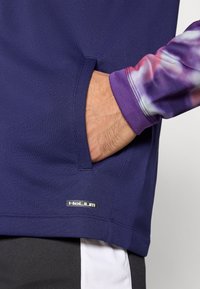 Lila sportos felső hosszú, batikolással mintázott ujjal, oldalzsebbel és a szegély közelében elhelyezett márkajelzéssel. Sima, rugalmas anyag.