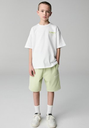 Bílá bavlněná tričko s zeleným logem "ALLSAINTS", doplněné světle zelenými kraťasy a bílými teniskami s černými akcenty. Jednoduchý design.