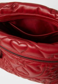 Červená kožená taška s prošívaným srdcovým designem. Obsahuje zipové uzavření a vnitřní kapsu. Hladký povrch s lesklým finišem.