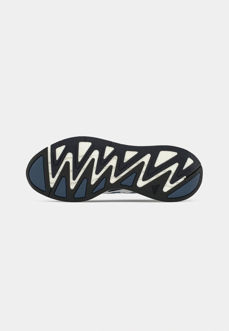 Semelle de sneaker noir et blanc avec des sections de grip bleu et un motif de treads en zigzag pour l'adhérence, photographiée sur un fond blanc.
