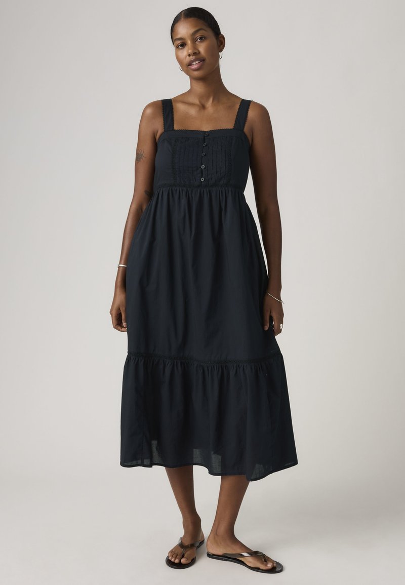 Femme portant une robe midi noire sans manches avec des boutons et des sandales, debout devant un fond clair uni.