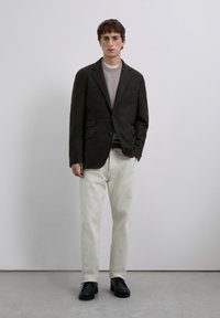 Joven de pie vestido con un blazer oscuro, un suéter gris claro, pantalones blancos y zapatos negros frente a un fondo gris claro liso.