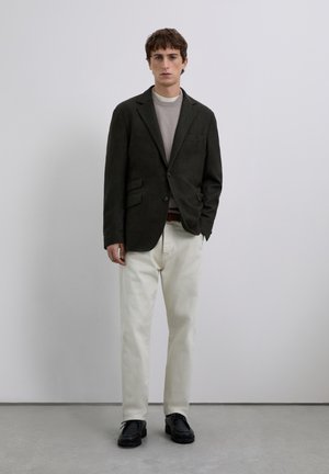 Joven de pie vestido con un blazer oscuro, un suéter gris claro, pantalones blancos y zapatos negros frente a un fondo gris claro liso.