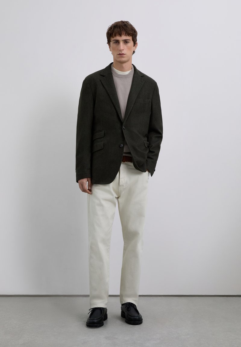 Joven de pie vestido con un blazer oscuro, un suéter gris claro, pantalones blancos y zapatos negros frente a un fondo gris claro liso.