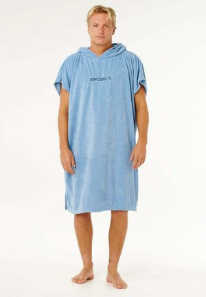 Mężczyzna stojący boso, ubrany w jasnoniebieski ponczo z kapturem i logo "RIP CURL" na jednolitym białym tle.