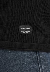 Črni pleteni pulover z teksturirano površino, ki ima pravokotno oznako z napisom "JACK&JONES" in "QUALITY ESSENTIALS SINCE 1990."