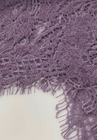 Gros plan d’un tissu en dentelle violet avec des motifs floraux et des bords délicatement effilochés sur un fond clair.