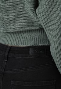 Green knitted sweater with a ribbed texture, paired with black denim jeans featuring a leather label marked "ONLY" on the back.