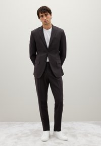 Completo grigio scuro da uomo con giacca su misura e pantaloni dritti, abbinato a una maglietta bianca e sneakers bianche. Texture liscia e design minimalista.