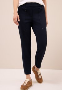 Donna che indossa pantaloni tapered blu navy con tasche a zip frontali e sneakers in suede marrone, che sta in piedi su un pavimento chiaro contro uno sfondo beige.