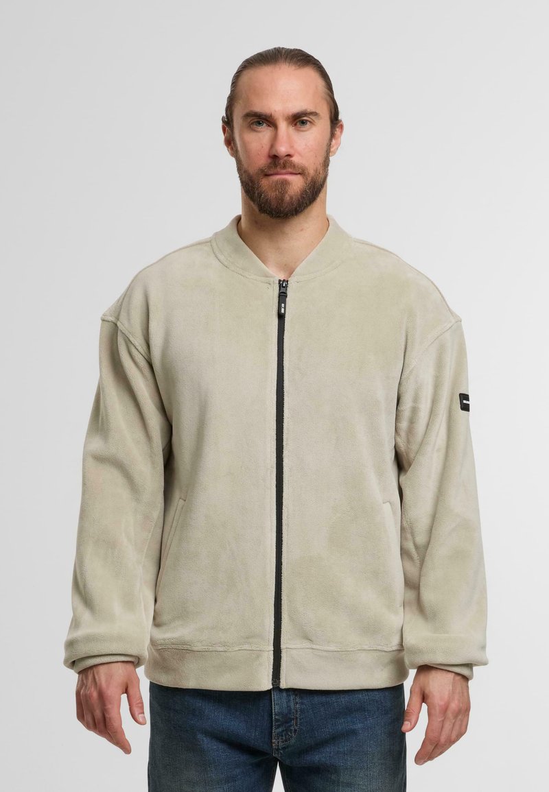 Lichtbeige zip-up jack gemaakt van zachte, gestructureerde stof, met een ronde halslijn en contrasterende zwarte rits. Iets oversized pasvorm.