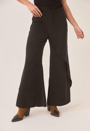 Mujer con pantalones negros de pierna ancha y detalle de envoltura asimétrica, combinados con botines marrones de tacón y punta afilada.