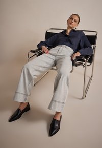 Lichtgrijze wijde jeans met zichtbare stiksels, gecombineerd met een donkerblauw overhemd met knopen en zwarte puntige loafers, zittend op een elegante zwarte stoel.