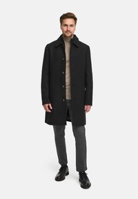 Svart ullfrakk med en strukturert krage og fem knapper, brukt over en beige genser med høy hals og grå bukser, kombinert med svarte sko.