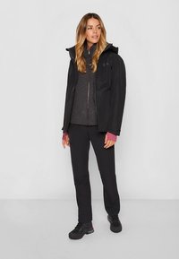 Svart vanntett jakke med hette, grå fleeceinnside, svarte bukser og mørke sko. Merkbar fargekontrast på ermene ved mansjettene.