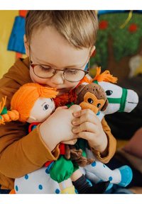 Kind met een bril omarmt meerdere knuffels, waaronder een bruine aap, een paard met groene accenten en een pop met oranje haar.