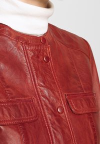 Röd läderjacka med en slät yta, knappar och två framfickor. Designen inkluderar en rundad krage och synliga sömnadsdetaljer.