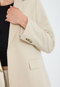 Blazer beige à un bouton, avec des revers crantés et trois boutons décoratifs sur la manche. Tissu lisse avec une coupe ajustée.