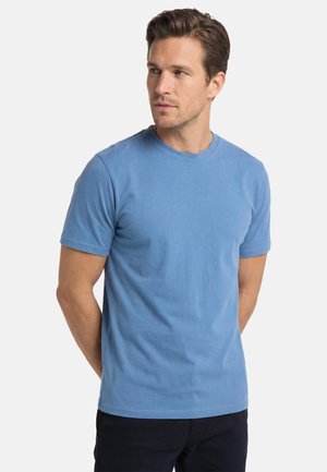 Mand med brunt hår iført en ensfarvet blå t-shirts med korte ærmer og sorte bukser, som kigger let mod venstre med hænderne bag ryggen.