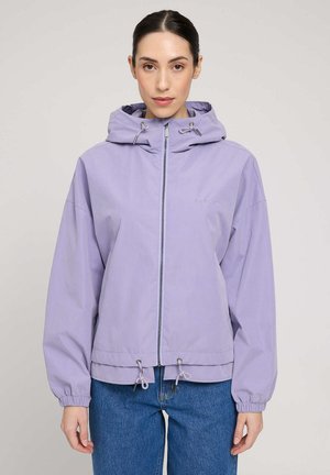 Frau trägt eine hellviolette Kapuzenjacke mit Reißverschluss und Kordeln und blaue Jeans, steht vor einem schlichten hellen Hintergrund.