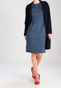 Robe bleu profond à manches courtes avec un motif texturé de feuilles, associée à un cardigan bleu marine foncé et des escarpins rouges.