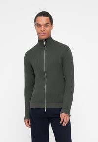 Sweater zippé en tricot vert foncé avec un col montant, des poignets et un ourlet côtelés. Motif texturé sur l'ensemble.