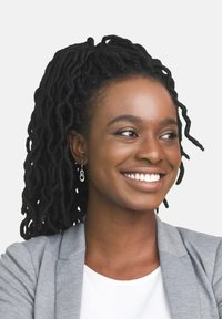 Porträtt av en kvinna med långt, mörkt lockigt hår, klädd i en ljusgrå kavaj över en vit topp och silverfärgade hoopsmycken, leende.