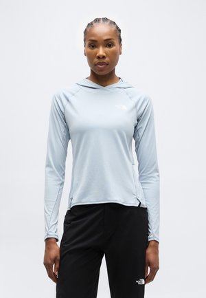 Kobieta z warkoczami, ubrana w jasnoniebieską bluzę z kapturem i długim rękawem oraz czarne spodnie, stojąca na tle jednolitego białego tła.