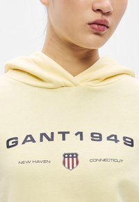 Žltá mikina s kapucňou s jemnou textúrou, zdobená potlačeným logom a textom "GANT1949" v tmavomodrej farbe, s erbom v tvare štítu a detailom "New Haven".