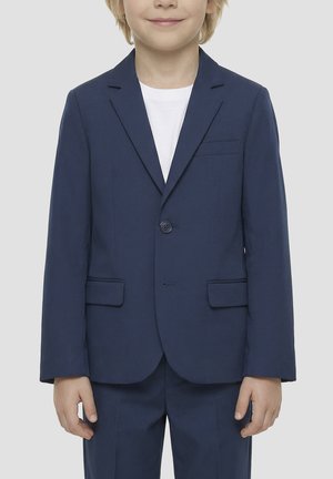 Kind draagt een marineblauw colbert en bijpassende broek over een wit overhemd, staat met ontspannen handen langs het lichaam.