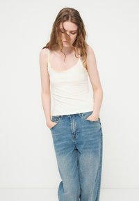 Jonge vrouw met golvend haar, die een witte mouwloze top en losse blauwe spijkerbroek draagt, staand met handen in de zakken tegen een effen achtergrond.