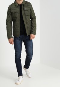 Gequilted olijfgroene jas met twee voorzakken, gedragen over een zwarte top, gecombineerd met donkere spijkerbroek en witte sneakers, staand op een lichte vloer.