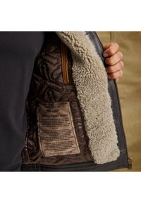 Vnútorné vyhotovenie hnedej koženej bundy má krémovo sfarbenú ovčiu srsť, prešívanú hnedú látku a béžovú starostlivostnú nálepku s čiernym textom.