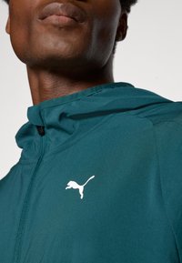 Teal zip-up hoodie med en texturerad finish, med en vit Puma-logotyp på vänster bröst och en hög krage för ökad täckning.