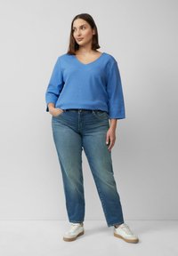 Lichtblauwe, gestructureerde trui met een V-hals en driekwart mouwen, gecombineerd met lichtgewassen denim jeans en witte sneakers met beige accenten.