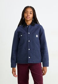 Una donna con lunghe trecce indossa una giacca blu navy con bottoni, dotata di tasche a toppa, e pantaloni di velluto a coste color bordeaux, in piedi su uno sfondo semplice.