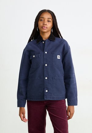 Una donna con lunghe trecce indossa una giacca blu navy con bottoni, dotata di tasche a toppa, e pantaloni di velluto a coste color bordeaux, in piedi su uno sfondo semplice.