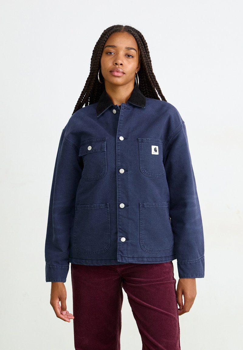 Una donna con lunghe trecce indossa una giacca blu navy con bottoni, dotata di tasche a toppa, e pantaloni di velluto a coste color bordeaux, in piedi su uno sfondo semplice.