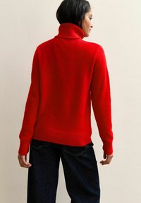 Maglione rosso in morbido knit con un alto collo a rolo e polsini a costine, abbinato a jeans neri a gamba larga. La texture appare liscia e calda.