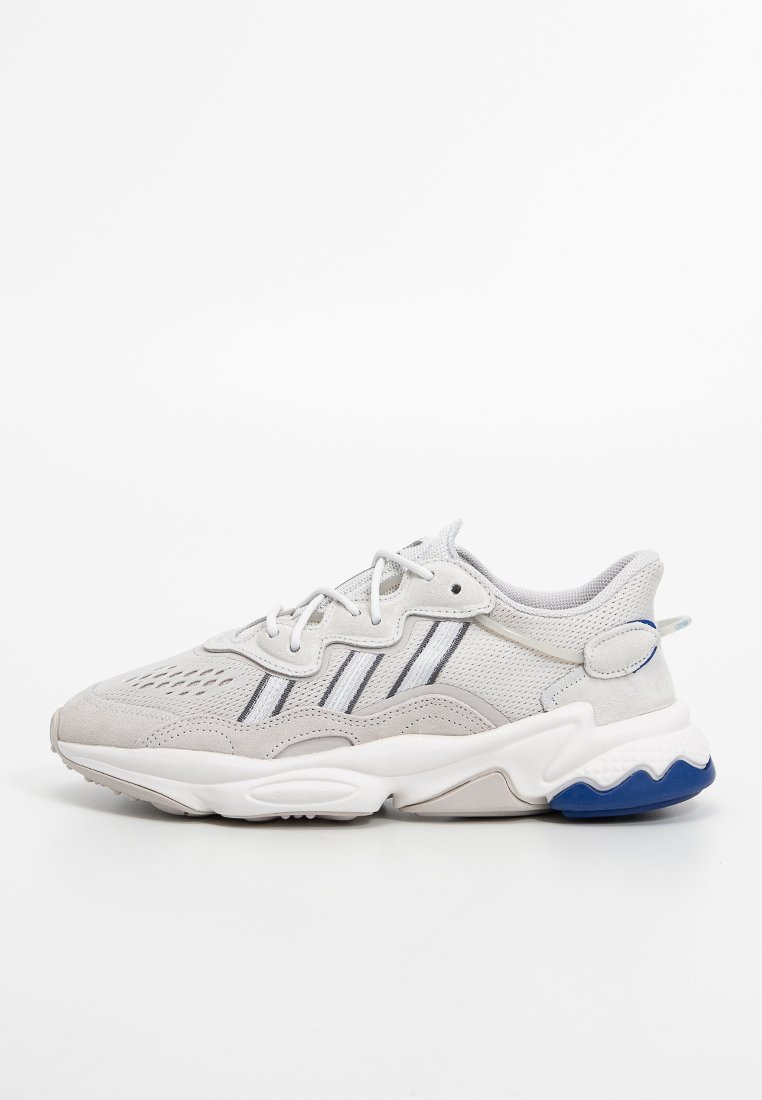 Zapatilla gris claro con paneles de malla, acentos de ante y tres franjas estrechas. Cuenta con una suela blanca gruesa con detalles en azul y cordones redondos.