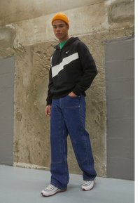 Čierna mikina s bielym pruhom, modré voľné džínsy a biele tenisky s červenými akcentmi. Svetložltý pletený klobúk dotvára vzhľad.