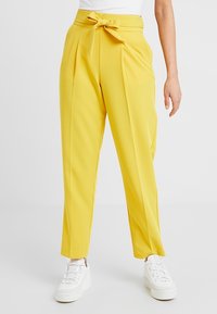 Geel, hoog-glepend broek met een strikriem, voorzien van plooien, taps toelopende pijpen en een gladde textuur. Gecombineerd met witte sneakers.