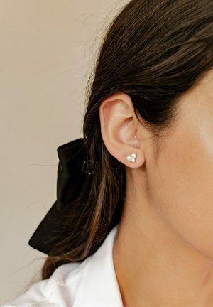 Silberne Ohrstecker mit zwei weißen Perlen und einem kleinen Kristall, am Ohr eines Modells befestigt. Die Haare sind mit einem schwarzen Band gestylt.