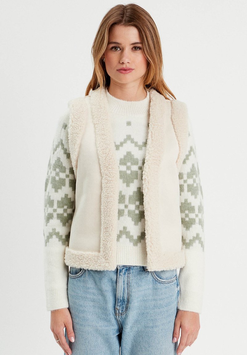 Cremefarbenes Weste mit flauschiger Textur, ausgestattet mit einem weichen Schererkragen. Darunter ein leichter Pullover mit grünem geometrischen Muster.